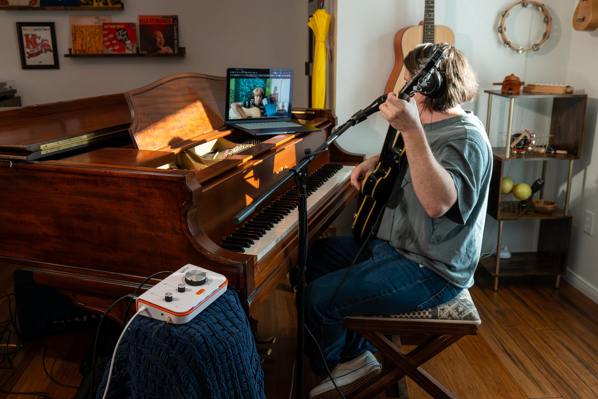 A man sits on a folding chair in a music room, playing a black and yellow acoustic guitar while singing into a microphone. He is wearing a gray t-shirt and blue jeans.